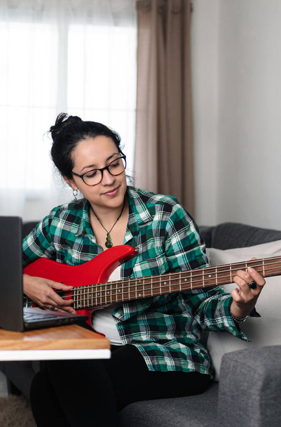 Brunette woman with glasses playing bass and using her computer at home. Vertical shot - Фото, изображение