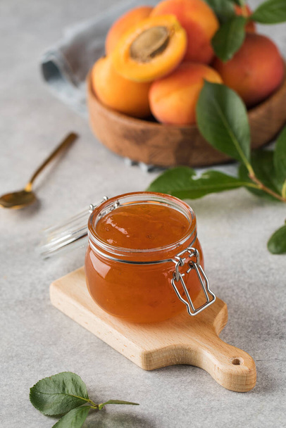 A jar of apricot jam on a light gray background, next to it is a golden spoon and a wooden plate with ripe apricots. The concept of fruit conservation. - Φωτογραφία, εικόνα