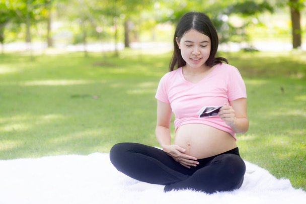 Happiness young asian woman pregnant sitting touch belly and looking photo with positive in the park, mother and health of pregnancy, happy of parenthood, one person, lifestyles and family concept. - Photo, Image