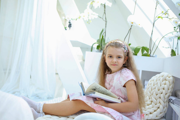 Banner with a cute little Caucasian girl in a pink dress with a book sitting in the children's room. Reading books. Learn to read. Education concept. Copy space on the left - Фото, изображение