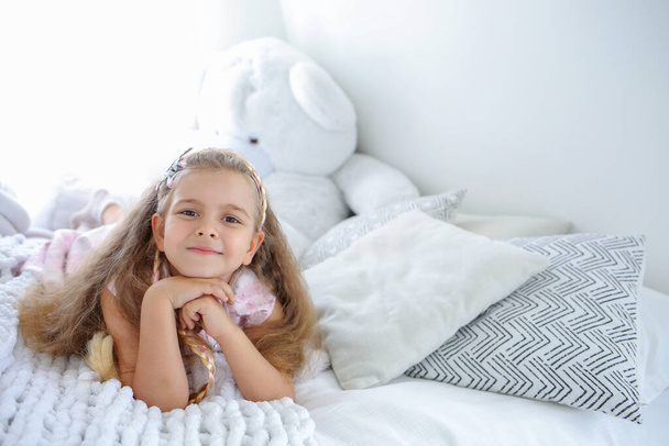 Transparent mit einem kleinen süßen Mädchen mit einem Lächeln liegt auf dem Bett. Helles Interieur im Kinderzimmer. Skandinavisches Interieur. Kindheitskonzept. Kopieren Sie das Leerzeichen rechts - Foto, Bild