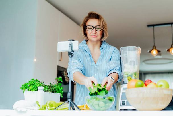 Middle age women is blogging for her wellness lifestyle channel about healthy balanced living in the kitchen of her home. Creating video content for social media with phone camera on a tripod. - Foto, Bild