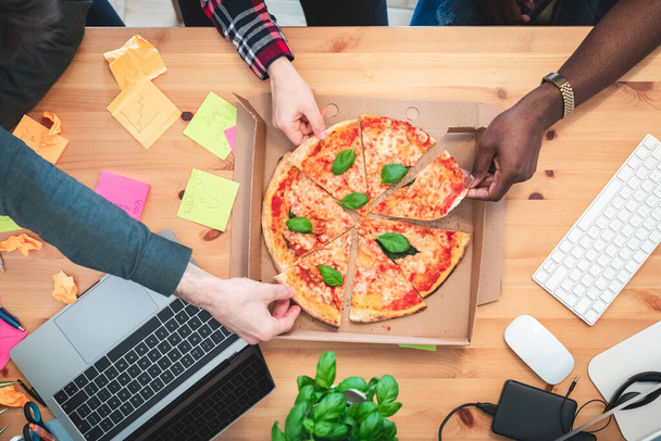Friends sharing pizza at startup home office or coworking space - Multiracial group of coworkers in small home office eating slices of pizza - Startup business or friends sharing a project - 写真・画像