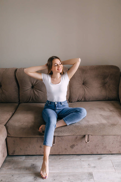 In cozy living room happy woman put hands behind head sitting leaned on couch - Фото, изображение