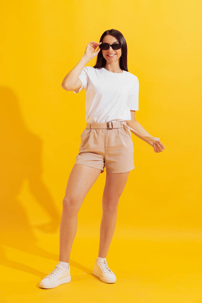 Summer fashion. Studio shot of young charming girl with long dark hair in shorts and t-shirt isolated on bright yellow background. Concept of beauty, art, youth, health. Copyspace for ad - Zdjęcie, obraz