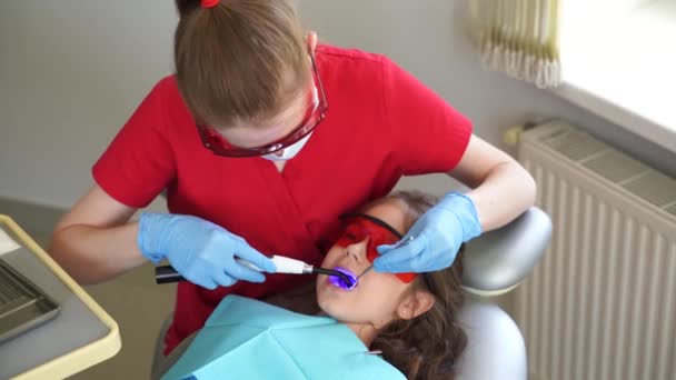 Uv illumination of photopolymer tooth filling procedure in dentistry. 4k video. Pediatric dentist in red protective glasses treats and removes caries in child girl - Imágenes, Vídeo