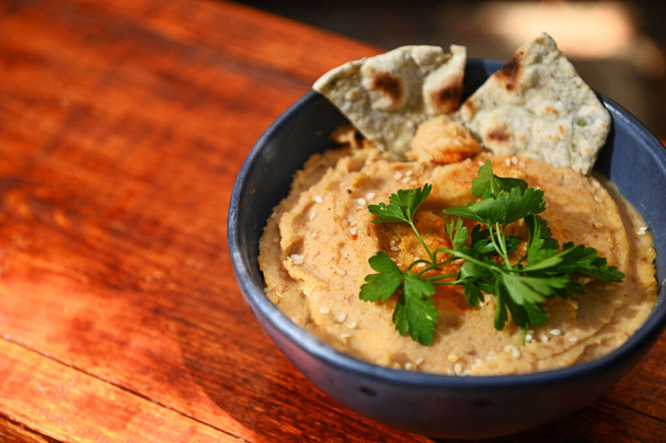 Close-up of a chickpea hummus dip with pita bread, parsley and sprinkled paprika on a blue ceramic bowl, on a rustic wooden table background with copy ad space for text. Still life. Food composition - 写真・画像