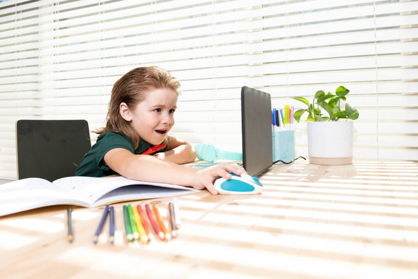 Fiksu koulupoika kotona tekemässä läksyjä. Pieni oppilas, jolla on kannettava tietokone valmiina opiskeluun. Koulun kaukosäädin, verkko-oppimista. Lapsi kotiluokassa oppiminen - Valokuva, kuva