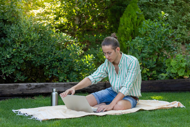 Young man with bun hairstyle and eyeglasses on opening his laptop to start working on his first online job sitting in the garden - Foto, Imagem