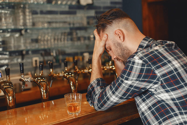 Man in a shirt holds a glass in his hands. Guy is sitting at the bar and holding his head. - Foto, Imagen