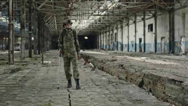 Wounded female soldier walking with crutch through destroyed factory after mission. Military woman in special forces uniform and helmet looking around abandoned building. - Πλάνα, βίντεο