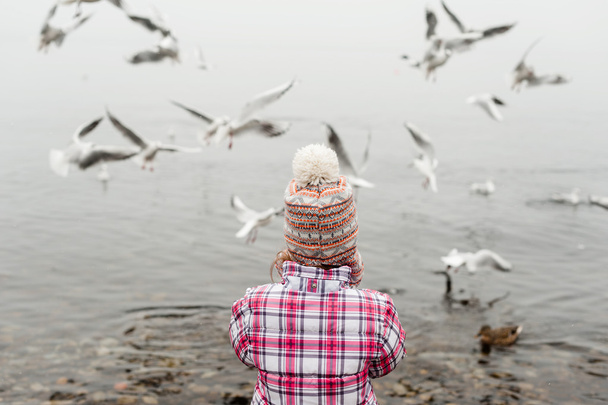 Malá holčička krmit ptáky na jezeře v zimě nosí teplé bundě a čepici - Fotografie, Obrázek