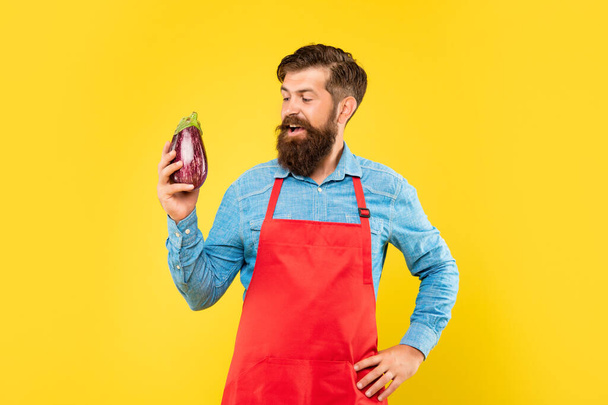 Happy man in red apron looking at eggplant yellow background, grocer. - Zdjęcie, obraz