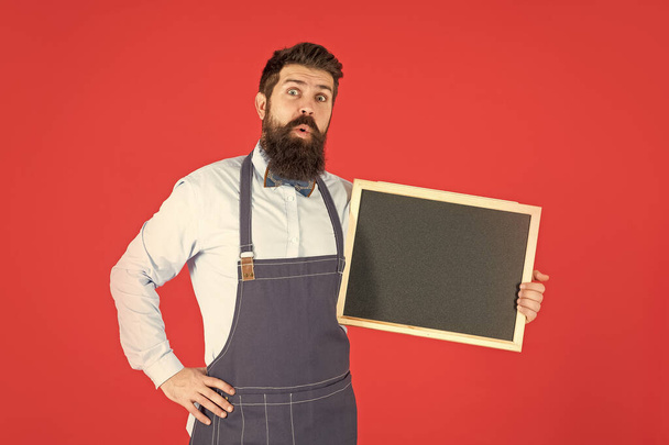 Surprised bearded man in barber apron hold school blackboard red background copy space, announcement board. - Photo, image