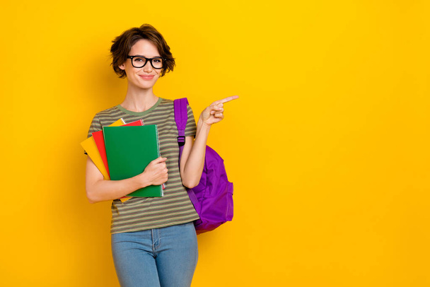 Photo of pretty sweet girl dressed green t-shirt eyewear rucksack holding book pointing empty space isolated yellow color background. - 写真・画像