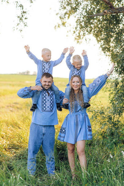 Happy parents carry their two twins sons on their shoulders and have a fun during walk on meadow. They are dressed in Ukrainian national embroidered shirts. - Foto, immagini