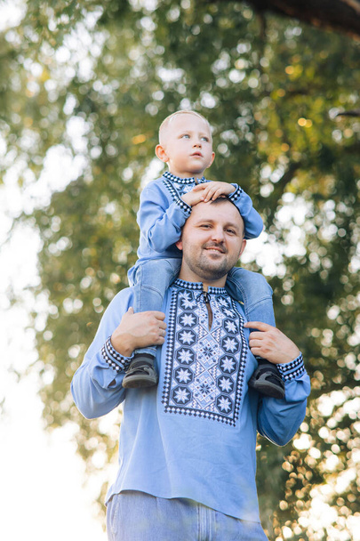 Happy father carries his son on his shoulders during walk on park. They are dressed in Ukrainian national embroidered shirts. - Foto, Imagen