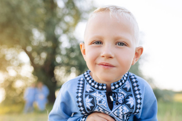 Happy child boy plays and lies on grass on meadow during walk. He is dressed in Ukrainian national embroidered shirt. - Foto, imagen