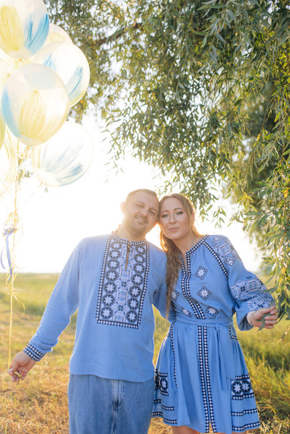 Happy couple smiles, embraces each other and holds air balloons during walk on meadow. They are dressed in Ukrainian national embroidered shirts. - Photo, image