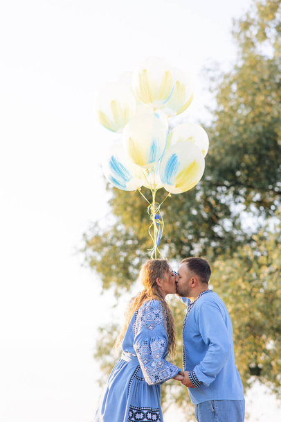 Happy couple kisses each other and holds air balloons during walk on meadow. They are dressed in Ukrainian national embroidered shirts. - Photo, image