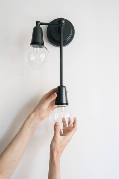 Vertical view of woman replace broken light bulb in black metal sconce. Electrician change energy saving lamp in apartment with white living room wall. Energy efficient equipment, household fixture - Valokuva, kuva