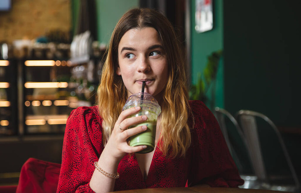 A young woman in a red dress in a cafe drinks a summer green drink ice latte. - Zdjęcie, obraz