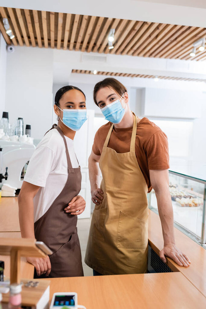 Multiethnic sellers in medical masks looking at camera in sweet shop  - 写真・画像