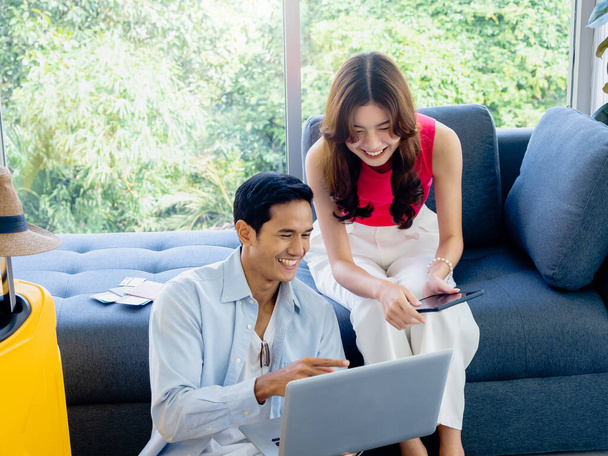 Happy Asian couple, young woman and man looking and using laptop computer and smartphone together for flight booking and trip information in living room near luggage, travel holiday summer vacation. - Foto, Imagen