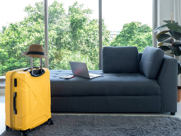Empty grey cozy fabric sofa seat with laptop computer and yellow suitcase with beach hat near huge glass window and green plant pot decoration in living room. Ready to summer vacation travel concept. - Photo, image