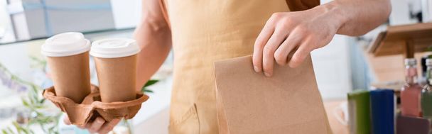Cropped view of salesman holding paper bag and coffee to go in confectionery, banner  - Foto, immagini