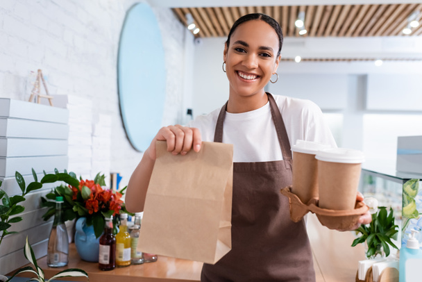Positive african american seller holding coffee to go and paper bag in sweet shop  - Foto, imagen