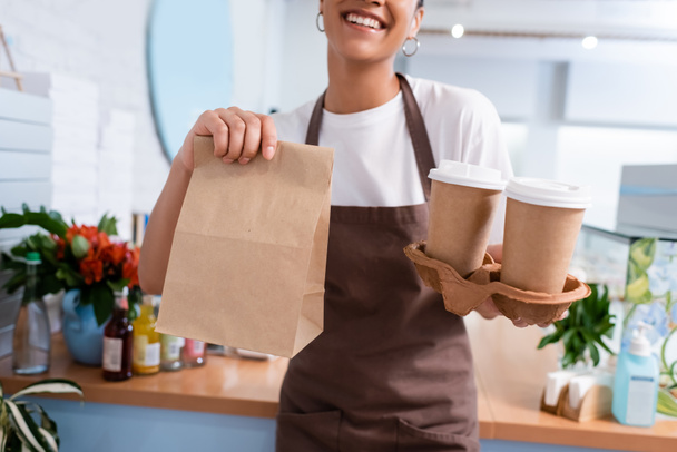 Cropped view of african american seller holding paper bag and takeaway coffee in confectionery  - Foto, imagen