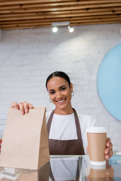 Positive african american seller holding paper cup and bag in sweet shop  - Zdjęcie, obraz