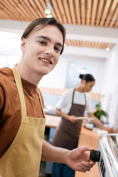 Smiling barista in apron looking at camera near coffee machine in sweet shop  - Photo, image