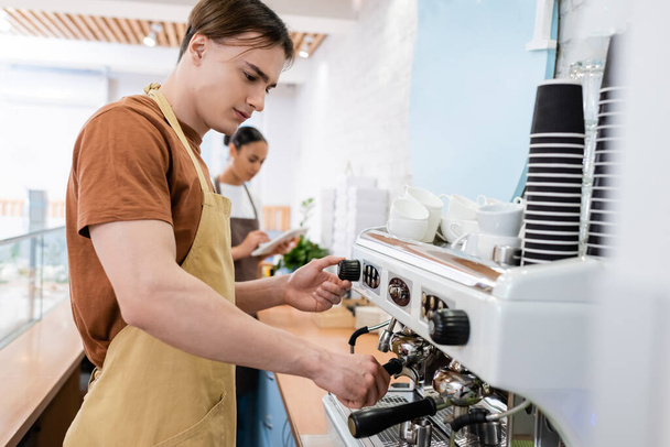 Young barista in apron using coffee machine near blurred african american saleswoman in confectionery  - 写真・画像