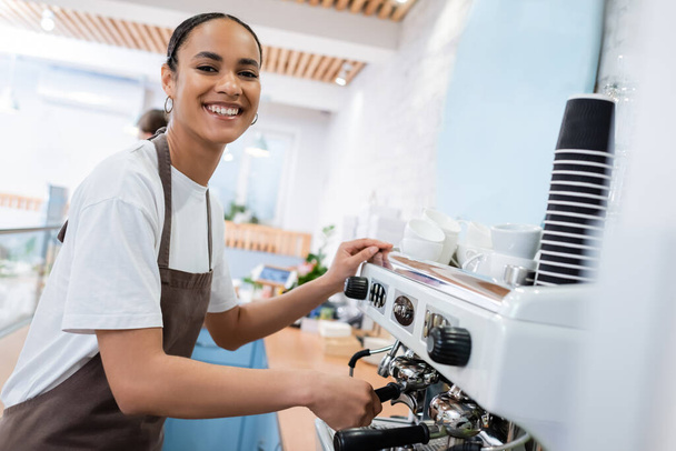 Cheerful african american barista making coffee and looking at camera in confectionery  - Фото, изображение