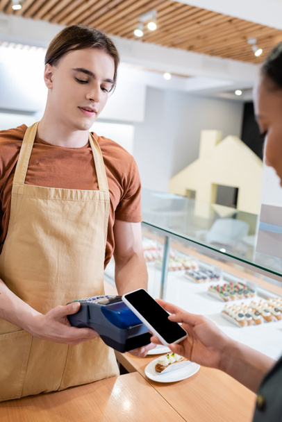 Seller holding payment terminal near african american customer with smartphone in sweet shop  - Фото, изображение