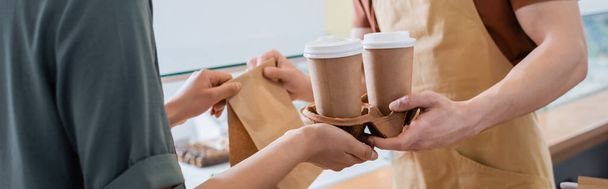 Cropped view of african american client taking coffee to go and paper bag from seller in sweet shop, banner  - Foto, Imagen