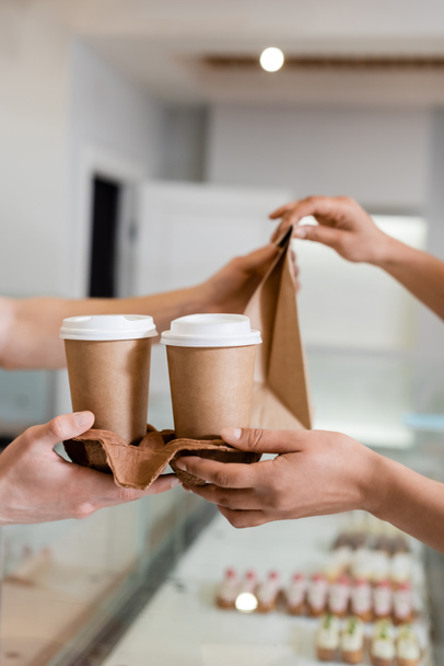 Cropped view of african american customer taking paper cups and bag from seller in sweet shop  - 写真・画像
