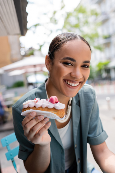 Positive african american client holding eclair with cream on terrace of confectionery  - Фото, зображення