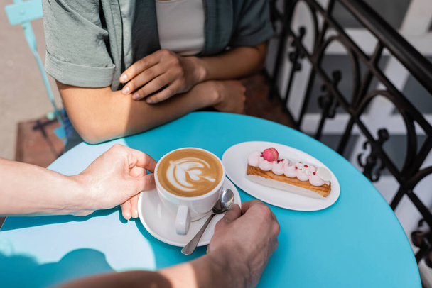 Cropped view of seller putting cappuccino on table near dessert and african american client on terrace of sweet shop   - Foto, Imagem