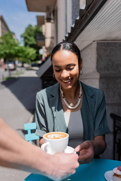 Smiling african american woman taking cappuccino from waiter near dessert on terrace of sweet shop  - Фото, зображення
