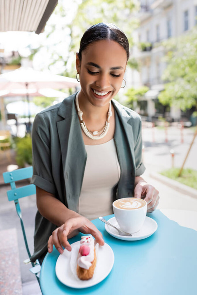 Smiling african american woman holding cappuccino and taking eclair on terrace of sweet shop  - Фото, зображення