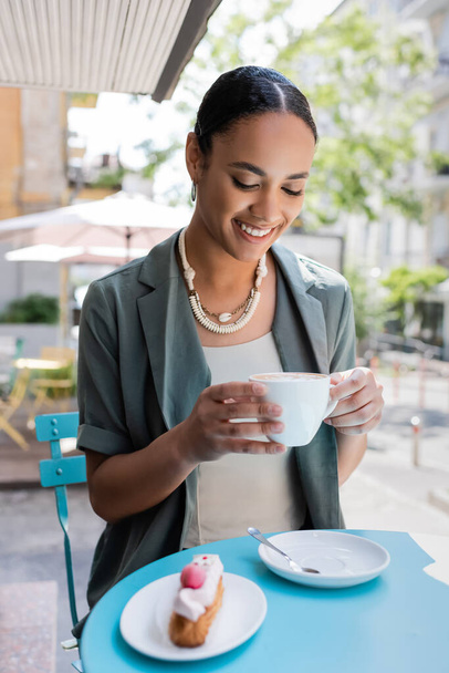 Positive african american woman holding cappuccino near blurred dessert on terrace of sweet shop  - Zdjęcie, obraz
