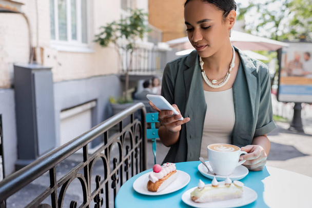 Afroamerikanerin benutzt Smartphone in der Nähe von Kaffee und Eclairs auf der Terrasse eines Süßwarengeschäfts  - Foto, Bild