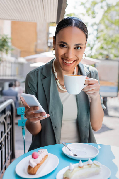Smiling african american client holding coffee and smartphone near eclairs on terrace of sweet shop  - Fotografie, Obrázek