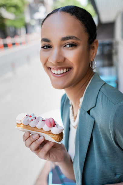 Pretty young customer holding delicious eclair and looking at camera on terrace of cafe  - Valokuva, kuva