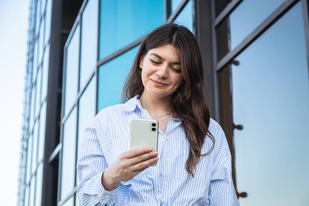 Beautiful business young woman with a smartphone on the background of a glass building. - Fotoğraf, Görsel
