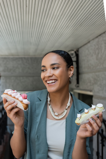 Cheerful young african american woman holding eclairs with cream near sweet shop  - Foto, afbeelding