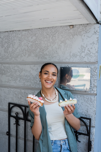 Cheerful african american woman holding sweet eclairs near confectionery  - Fotó, kép